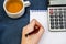 Pen in female hand, notebook, calculator and cup of tea on blue background