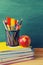 Pencils, rulers, scissors in a glass, a red apple, a stack of notebooks and textbooks on the background of a green school board