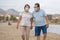 Pensioner woman and her husband taking romantic walk together - happy retired mature couple in face mask walking on the beach in