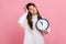 Pensive confused female with dark hair holding big wall clock, looking away, keeping hand on head.