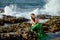 Pensive girl in a green mermaid costume sits on the rocks on the seashore on the background of water splashes and looks to the sid