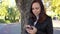 Pensive girl with smartphone in hands in city park. woman is looking at the screen of mobile phone against background of green tre