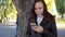 Pensive girl with smartphone in hands in city park. woman is looking at the screen of mobile phone against background of green tre