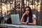 Pensive hipster girl in white shirt looking at camera while sitting on a wooden bench with an open laptop in the