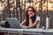 Pensive hipster girl in white shirt looking at camera while sitting on a wooden bench with an open laptop in the