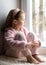 Pensive kid sitting on windowsill at home looks out window to snow