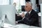 Pensive mature businessman holding glasses and looking at computer monitor near multicultural colleagues in office