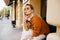 Pensive pale girl in vintage brown sweater touching chin with fingers and looking up. Outdoor portrait of stylish lady