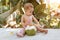 Pensive and passionate infant baby sits on the ground and eats and drinks green young coconut with spoon and drinking straw