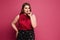 Pensive plus-size model girl with bright makeup and with stylish hairstyle in a red blouse and skirt posing at the pink