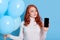 Pensive red haired European woman thinks celebrate birthday holiday party, holds modern mobile phone with blank screen and bunch