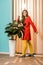 pensive retro styled woman standing at ficus plant in flowerpot at colorful apartment doll