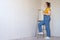 A pensive woman with brush and ladder looks dreamily in a room with an unpainted wall with copy space. Independent