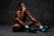 Pensive young African-American basketball player in sportswear sitting on a floor with a ball over dark background.