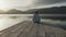 Pensive young woman with long hair seen from behind looks at the lake at sunset sitting on an old wooden pier