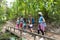 People Group With Backpacks Walking On Bridge Trekking On Forest Path, Young Men And Woman On Hike