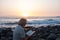 People senior with white hair and beard sitting on the beach of pebbles reading a book. Waves of the ocean. Sunset on background