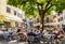 People sit on the open veranda of a cafe in the shade of a large tree on the street of a Malcesine town