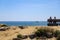 People sitting on a wooden benches overlooking the vast blue ocean water surrounded by lush green plants and rocks