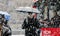 People under umbrellas crossing snowy city streetl and a driving bus behind them