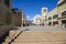 People walking through a courtyard surrounded by apartments,shops and tall brown light posts with mountains, clouds and a clear