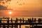 People Walking on Sunset over Bridge. Backlight Shot. Silhouettes of Men, Women, Children and Dog in Front of Lake.