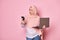 Perplexed Arab Muslim pregnant woman in hijab, looking at her smartphone, posing with laptop over pink color background