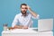 Perplexed young bearded man in light shirt sit and work at desk with pc laptop isolated on pastel blue background