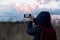 A person in a blue hooded jacket and with a red backpack takes pictures of nature,the sky and clouds on the phone