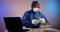 A person in blue protective clothing with a hood, gloves, a respirator and orange glasses is sitting at a table with a