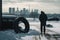 person, changing tires in snowy parking lot, with view of the city in the background