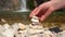 Person during a halt on the bank of mountain river on hiking trip collects pyramid of stones, close up, waterfall in