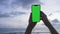 Person hands hold vertical mobile phone with green screen against background ocean and clouds sky. Use green screen for