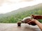 Person hands holding ceramic teapot pouring hot herbal tea in small cup on wooden table on natural green mountain view.