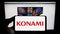 Person holding cellphone with logo of Japanese company Konami Group Corporation on screen in front of business webpage.