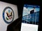 Person holding cellphone with web page of United States Department of State (DOS) on screen in front of logo.