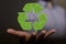 Person holding an illustrative green recycle symbol in his palm with a blurry background.