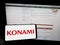 Person holding mobile phone with logo of Japanese company Konami Group Corporation on screen in front of web page.