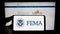 Person holding mobile phone with seal of Federal Emergency Management Agency (FEMA) on screen in front of web page.