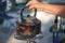 A PERSON PLACING A BLACKENED WATER KETTLE ONTO A GAS PLATE