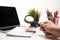 Person`s hand holding a magnifier on a desk having a laptop, calculator, organizer, crayons, notepad and a green plant