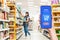 A person's hand holds a mobile phone, with a shopping basket on the screen. In the background, a worker selects products in a