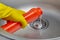 Person's hand in a yellow rubber glove pours pipe cleaner down the drain of a metal kitchen sink