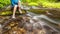 Person sits on the stone covered with moss in the center of rapid flow of the river, holding his feet in clear water