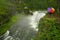 Person Standing in Rain with Rainbow Umbrella on Platform Looking at River Waterfall Pine Forest