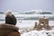 person in a winter hat admiring a sandcastle on a snowy beach with waves in the background