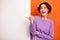 Photo of adorable excited girl dressed purple pullover pointing thumb white placard empty space isolated orange color