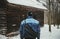 Photo of adventurer hiker in snow covered forest in front of abandoned cottage view from his back. Hiker Man in blue jacket