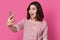 Photo of amazed surprised female holds mobile phone for making selfie portrait, has new image, bright pink makeup, dressed in