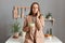 Photo of astonished sad upset brown haired woman wearing beige suit standing near table on kitchen at home, having breakfast or
